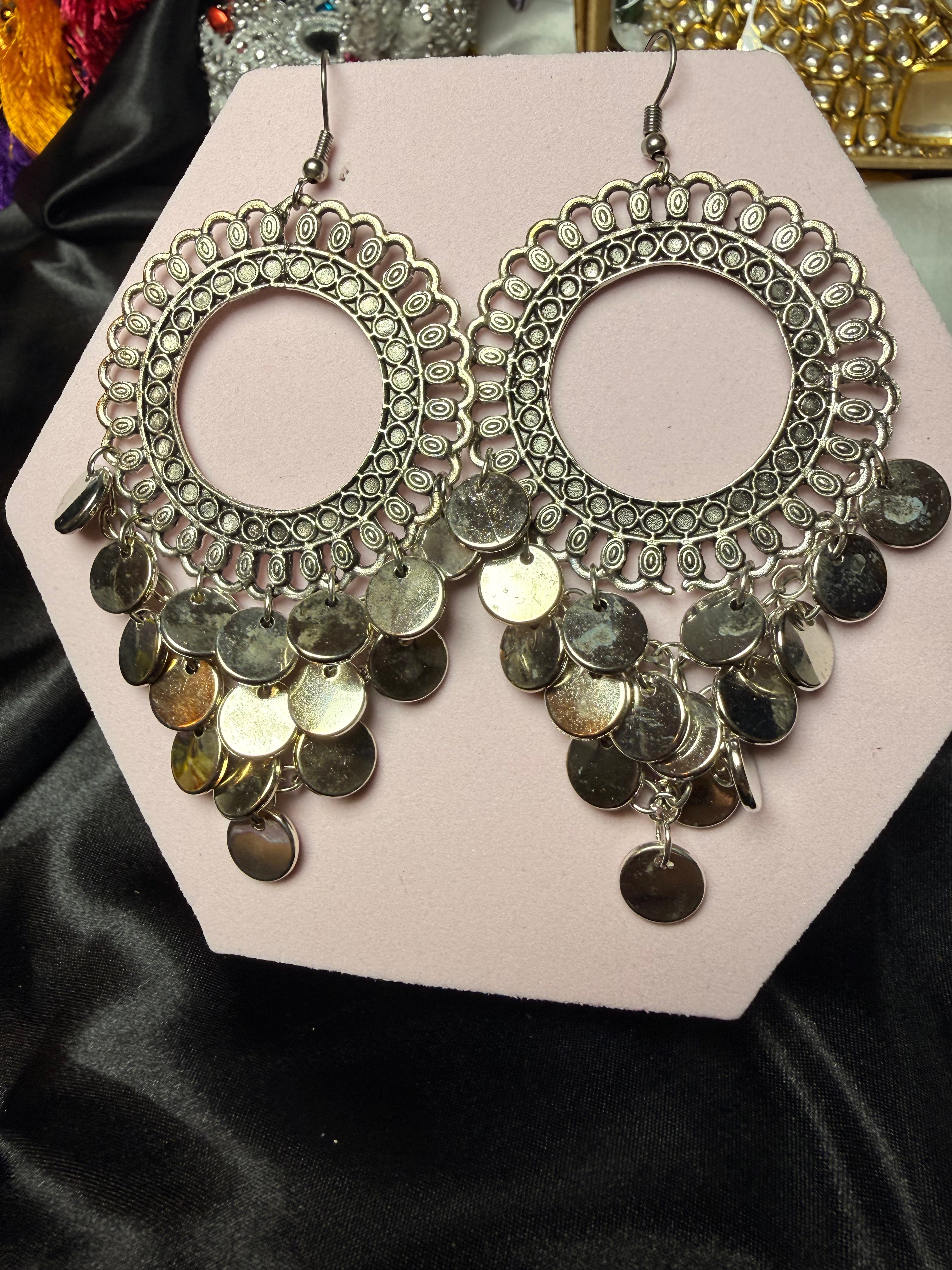 Pair of ornate earrings with coin details on a pink card against a black satin background.