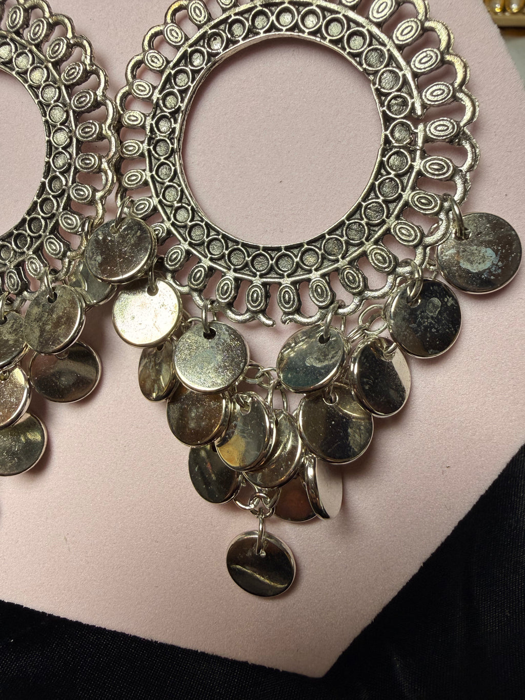 Ornate silver earrings with circular design and hanging coins on a pink stand.